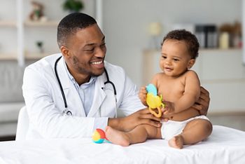 Portrait Of Smiling Black Doctor Making Medical Check Up For Little Baby Patient In Clinic, Handsome Young African American Pediatrist Looking At Adorable Infant Child In Diaper, Closeup Shot