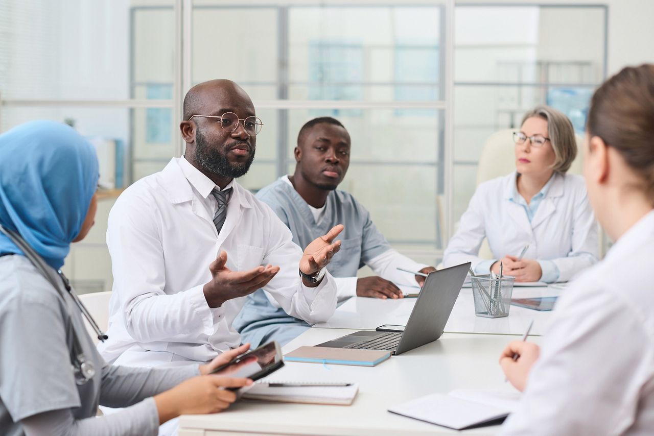 African American doctor in white coat sitting at table with laptop and discussing the way of treatment with other colleagues at meeting