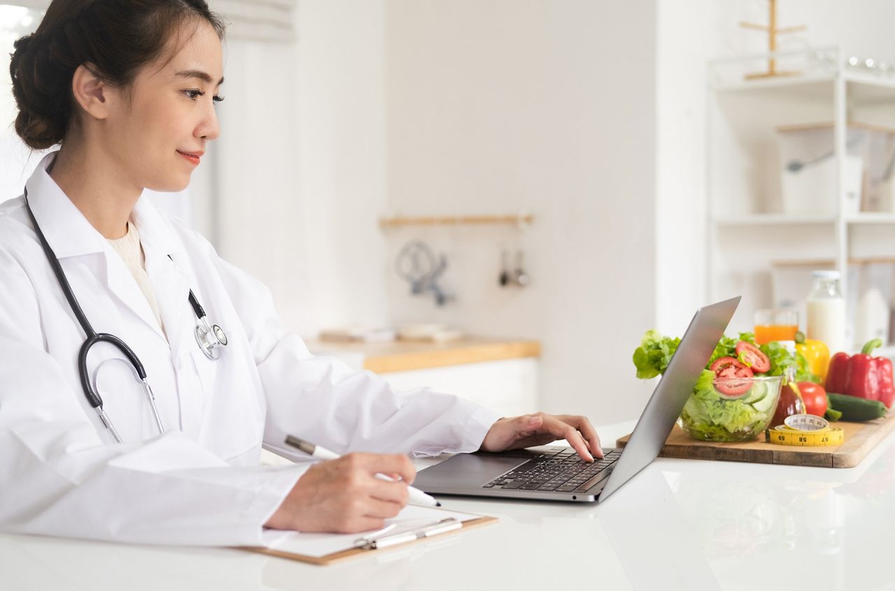 Close-up of dietitian hand writing with laptop giving online weight loss and healthy diet consultation with fresh vegetable fruit measuring tape and glass of clean water on desk against