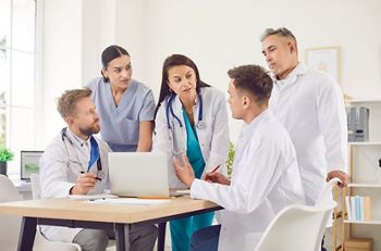 Group of professional doctors sitting with laptop at the desk on meeting discussing diagnosis, checking test results or planning treatment in clinic. Medical staff, healthcare and teamwork concept.
