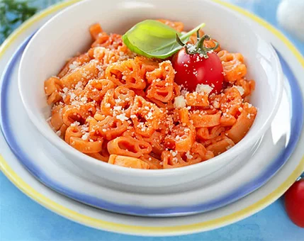 A white bowl filled with pasta in a rich tomato sauce, topped with a fresh basil leaf and a cherry tomato. The dish is sprinkled with grated cheese, adding texture and flavor. The setting features a bright blue background with hints of fresh ingredients nearby.