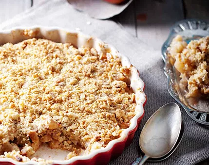 A freshly baked apple crumble served in a red ceramic pie dish, accompanied by a portion on a glass plate. The setting features a rustic wooden table with a silver spoon placed nearby. The crumble topping is golden and textured, highlighting the dessert's homemade appeal.