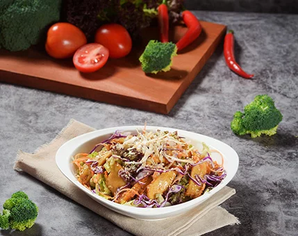 A colorful vegetable salad served in a white bowl, featuring broccoli, shredded cabbage, and tomatoes. The dish is garnished with grated cheese and surrounded by fresh produce, including chili peppers and leafy greens. The setting is a rustic gray tabletop with a wooden cutting board in the background.