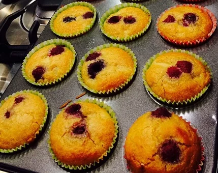 A batch of golden raspberry muffins is displayed on a baking tray, with colorful paper liners adding a vibrant touch. The setting appears to be a home kitchen, with a stovetop visible in the background. The muffins are evenly baked, with raspberries peeking through the tops, creating a warm and inviting visual.