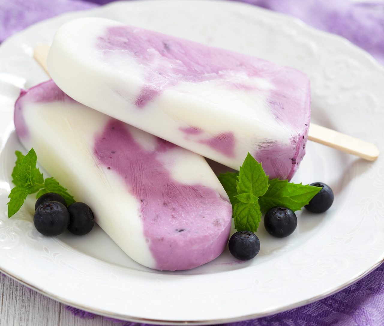 Zwei Joghurt-Blaubeer-Eis am Stiel liegen auf einem weißen Teller mit frischen Blaubeeren und Minzblättern.
