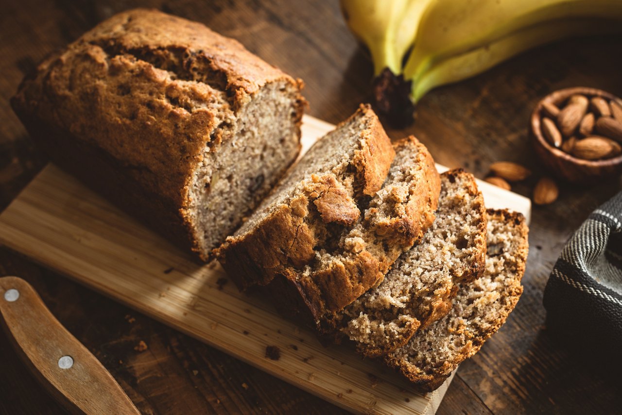 Bananenbrot in Scheiben auf einem Holzbrett, dahinter liegen reife Bananen und eine Schale mit Mandeln.