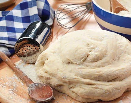 A close-up of freshly prepared dough resting on a wooden surface, surrounded by baking tools including a whisk, wooden spoon, and flour sifter. A blue and white checkered cloth adds a homely touch to the scene. The setting suggests a cozy kitchen environment ideal for baking activities.