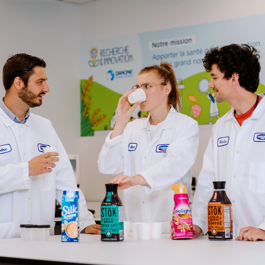 Trois employés de Danone en blouse de laboratoire se réunissent autour d'une table pour déguster et discuter de divers produits Danone, notamment Silk, Stok et International Delight.