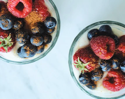Two glass bowls filled with a creamy parfait are topped with fresh raspberries, blueberries, and a sprinkle of cinnamon. The setting is bright and minimal, with a marble surface underneath. The vibrant colors of the berries contrast beautifully with the light cream base and warm cinnamon powder.