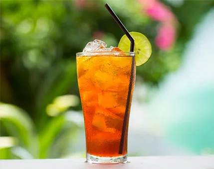 A tall glass of iced tea is served with ice cubes and a slice of lime on the rim. The drink is accompanied by a black straw and is set against a vibrant outdoor background with lush greenery and pink flowers. The scene evokes a tropical and refreshing mood, perfect for warm weather.