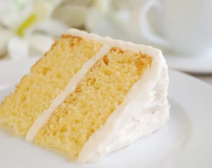 A close-up of a two-layer vanilla cake slice with creamy white frosting, served on a white plate. The setting includes soft floral accents and a blurred teacup in the background, suggesting a relaxed and elegant atmosphere. The cake's texture appears moist and fluffy, complemented by the smooth frosting.