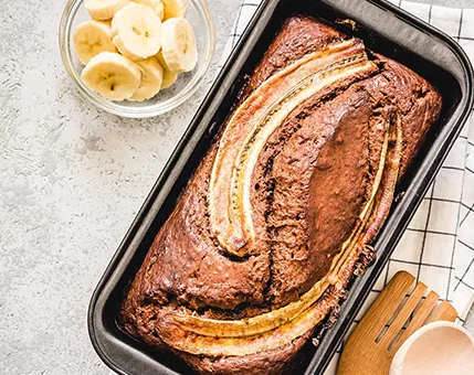 A loaf of banana bread is displayed in a black baking pan, topped with sliced bananas. The setting includes a bowl of fresh banana slices and wooden utensils on a light textured surface. The warm tones of the bread contrast with the neutral background, creating a cozy and inviting mood.