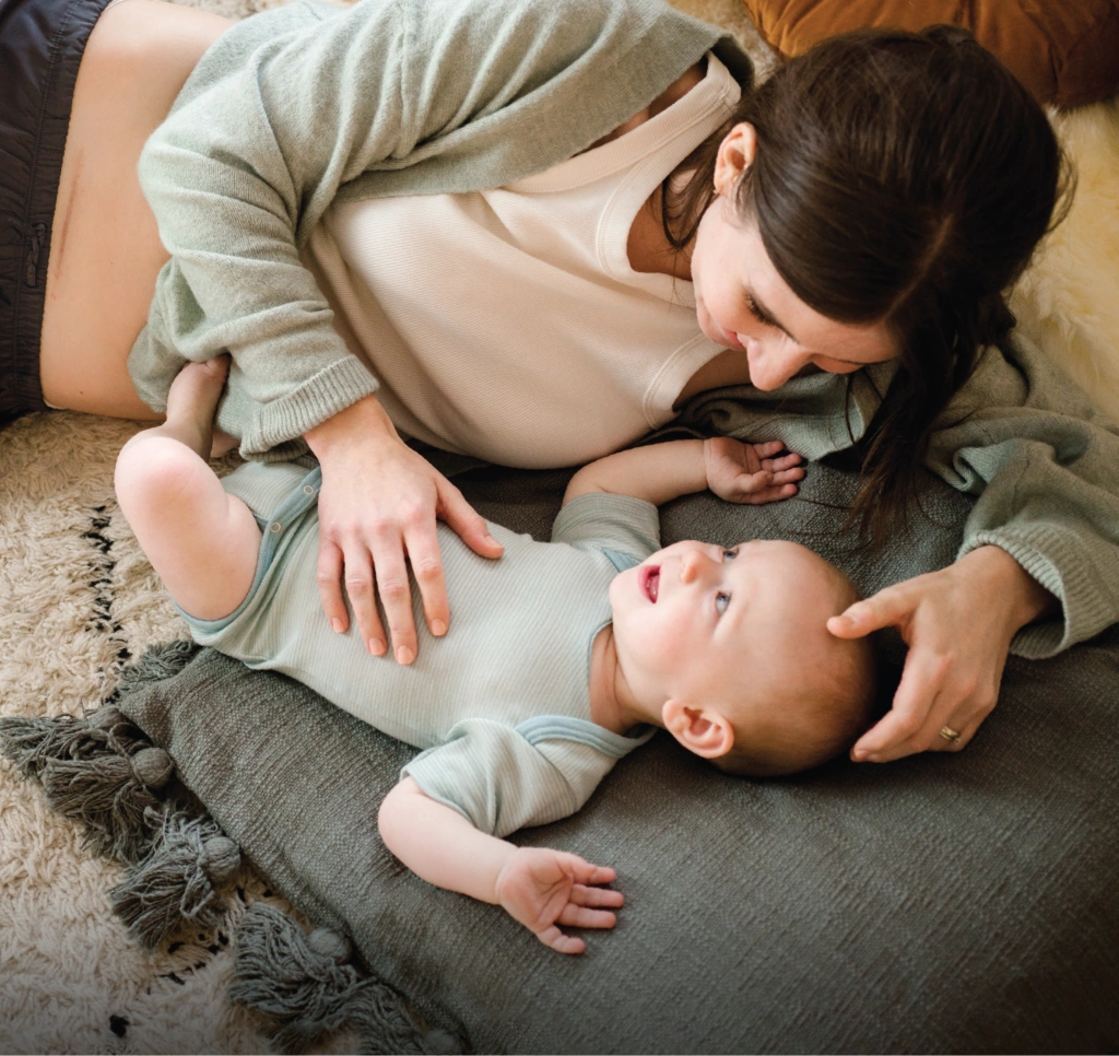A woman and a baby are lying together on a textured blanket, sharing a tender moment. The setting appears to be indoors, with soft natural light and neutral tones creating a warm, intimate atmosphere. The woman gently touches the baby's head, emphasizing nurturing and connection. The visuals include comfortable clothing and a plush rug, enhancing the sense of relaxation and care.