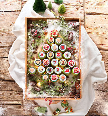 Un plateau en bois contient un arrangement creatif de cupcakes decores comme un calendrier de l'Avent, formant un sapin de No�l. Le decor comprend un fond en bois rustique enneige avec des arbres miniatures et des pommes de pin. Chaque cupcake est orne des chiffres 1 a 24 et de motifs festifs tels que des flocons de neige et des fleurs, ajoutant une touche de charme aux fetes.
