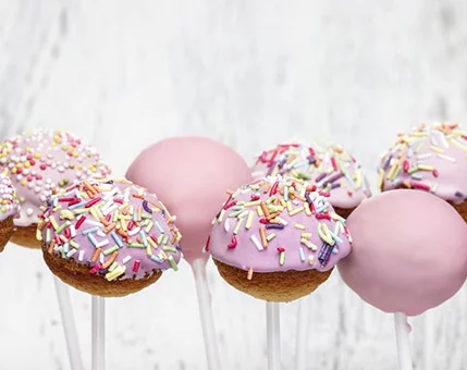A close-up of pink cake pops coated in icing and decorated with vibrant sprinkles. The treats are displayed on white sticks against a rustic white wooden background. The playful colors and textures evoke a cheerful and festive mood.