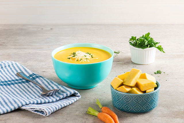 Un plato de crema de zapallo con zanahoria servido en un bowl azul, acompañado de crutones amarillos en un recipiente decorativo y perejil fresco en un fondo claro 