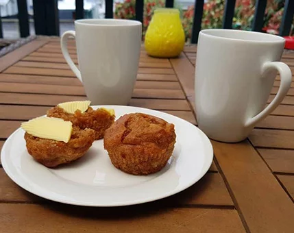 Two white coffee mugs are placed on a wooden patio table alongside a plate with two muffins topped with butter slices. A yellow decorative object is visible in the background, adding a pop of color to the setting. The scene suggests a casual outdoor breakfast or snack moment.