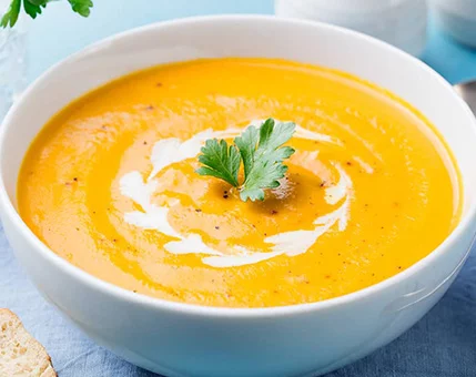 A bowl of vibrant orange carrot soup served in a white dish. The soup is topped with a swirl of cream and a fresh parsley leaf for garnish. The setting includes a light blue tablecloth and bread slices on the side, creating a cozy dining atmosphere.