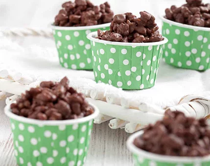 Small servings of chocolate-covered snacks are presented in green polka dot paper cups. The cups are arranged on a white surface with a woven tray in the background. The image evokes a casual and inviting mood, perfect for parties or gatherings.