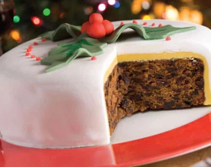 A traditional fruitcake topped with white icing and decorated with holly-shaped fondant and red berries. The cake is partially sliced to reveal its rich, dense texture filled with dried fruits. Set against a warm, festive background with blurred holiday lights.