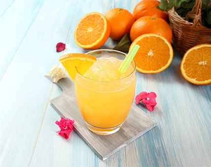 A glass of freshly prepared orange juice is served with a slice of orange and a green straw. The setting features a light blue wooden surface, surrounded by whole and sliced oranges in a wicker basket. Bright floral accents and vibrant colors enhance the summery mood.