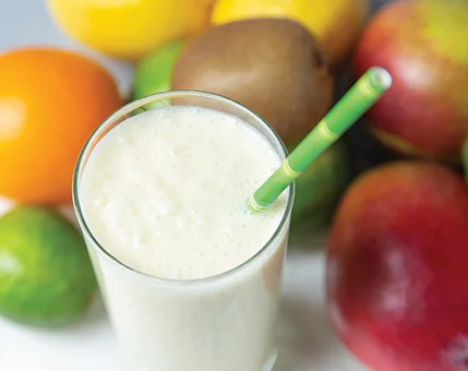 A creamy smoothie served in a clear glass with a green straw, surrounded by vibrant tropical fruits including mangoes, oranges, kiwis, and limes. The setting emphasizes freshness and healthy living. The bright colors of the fruits create a lively and appetizing visual appeal.
