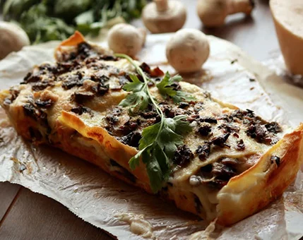 A freshly baked lasagna topped with melted cheese, mushrooms, and parsley garnish. The dish is presented on parchment paper with raw mushrooms and fresh herbs in the background. Warm lighting enhances the rustic and appetizing appeal of the meal.