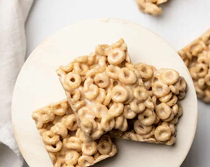 Close-up of honey-coated cereal bars stacked on a round white plate. The bars feature circular cereal pieces bound together, creating a textured and glossy appearance. A soft white cloth is partially visible in the background, adding a neutral and clean setting.