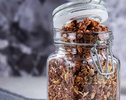 A clear glass jar with a hinged lid is filled with golden-brown granola, featuring visible nuts and seeds. The jar is placed on a textured surface with a blurred gray background, creating a rustic and cozy visual. The granola appears fresh and ready for serving or storage.