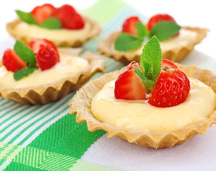 Four small tarts filled with creamy custard are topped with fresh strawberries and mint leaves. They are placed on a green and white striped cloth, creating a vibrant and appetizing presentation. The image highlights the dessert's rich texture and colorful garnish.