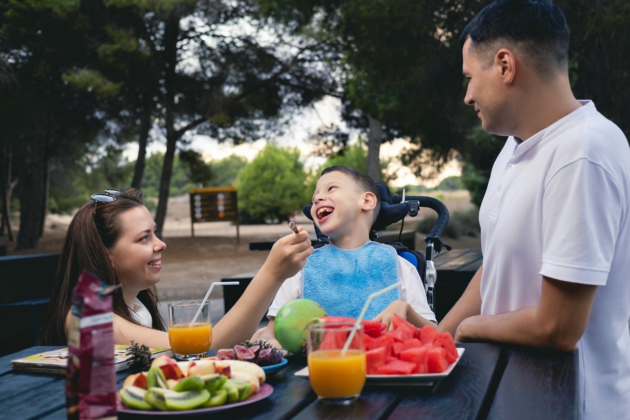 Loving parents his little 9s son with cerebral palsy enjoy picnic on nature. Married couple 35s wife and husband feeding his cute disabled son seated on wheelchair outdoor