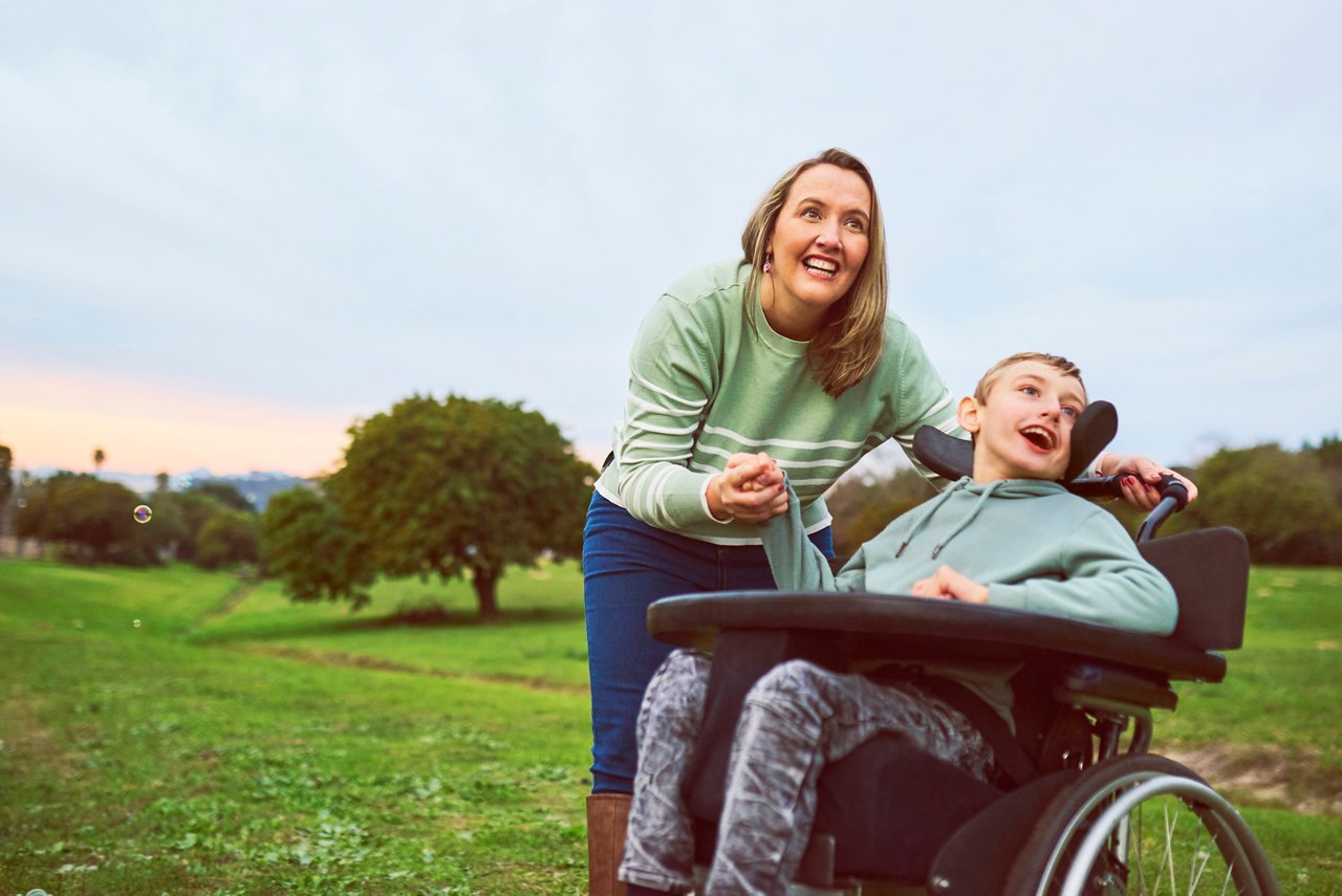 Woman and young special son sitting on wheelchair out for morning exercise, looking surprised and excited into distance, smiling cheerfully on open field of greeneries
