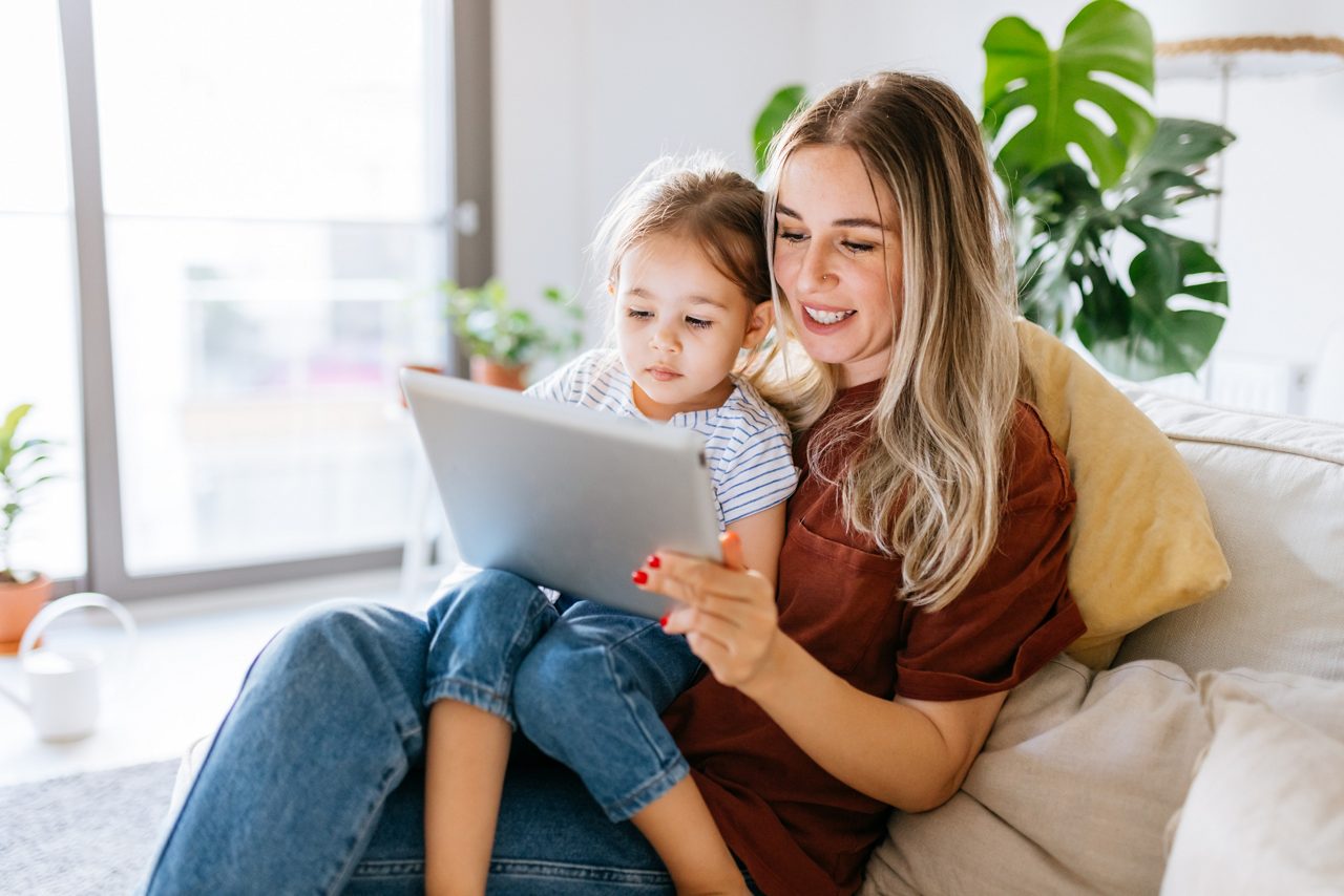 Mother and daughter using a digital tablet together. They are sitting on the sofa at home. The daughter is sitting on his mothers lap. Very hapy and smiling. Close up with Tight crop