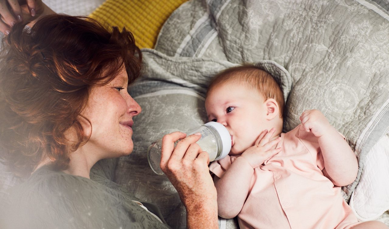 Eine Frau füttert ein Baby im rosa Strampler mit der Flasche auf einem Bett mit gemustertem Bettbezug.