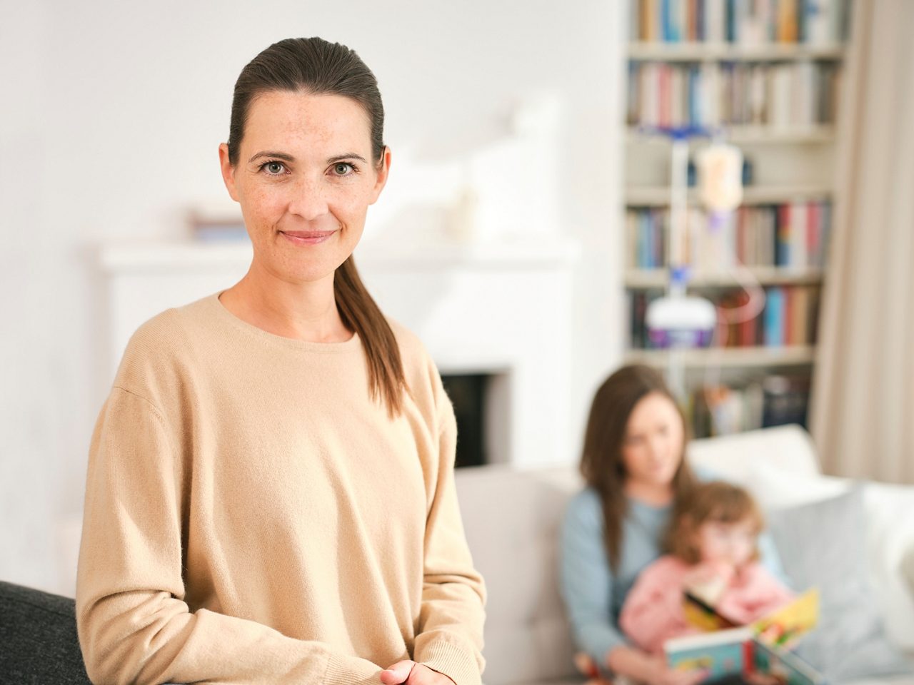 Eine Frau mit braunen Haaren steht im Vordergrund, im Hintergrund liest eine Frau einem Kind auf dem Sofa ein Buch vor.
