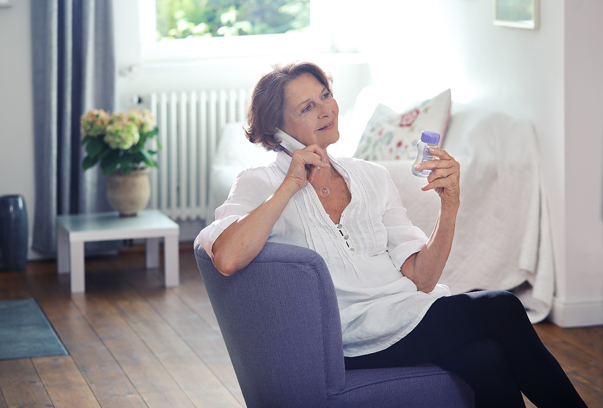 Eine ältere Frau spricht in einem Wohnzimmer am Telefon und hält eine Flasche mit lila Verschluss in der Hand.