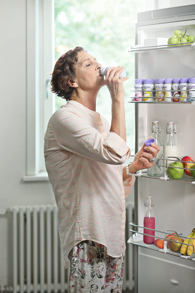 Frau steht neben Kühlschrank und trinkt Fortimel Trinknahrung