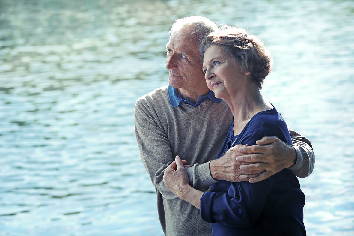 Mann hält seine an Krebs erkrankte Frau am See im Arm und sie blicken in die Ferne.