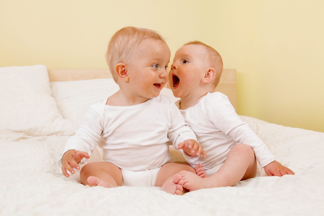 Two babies sitting on bed. One shouting in the other's ear.