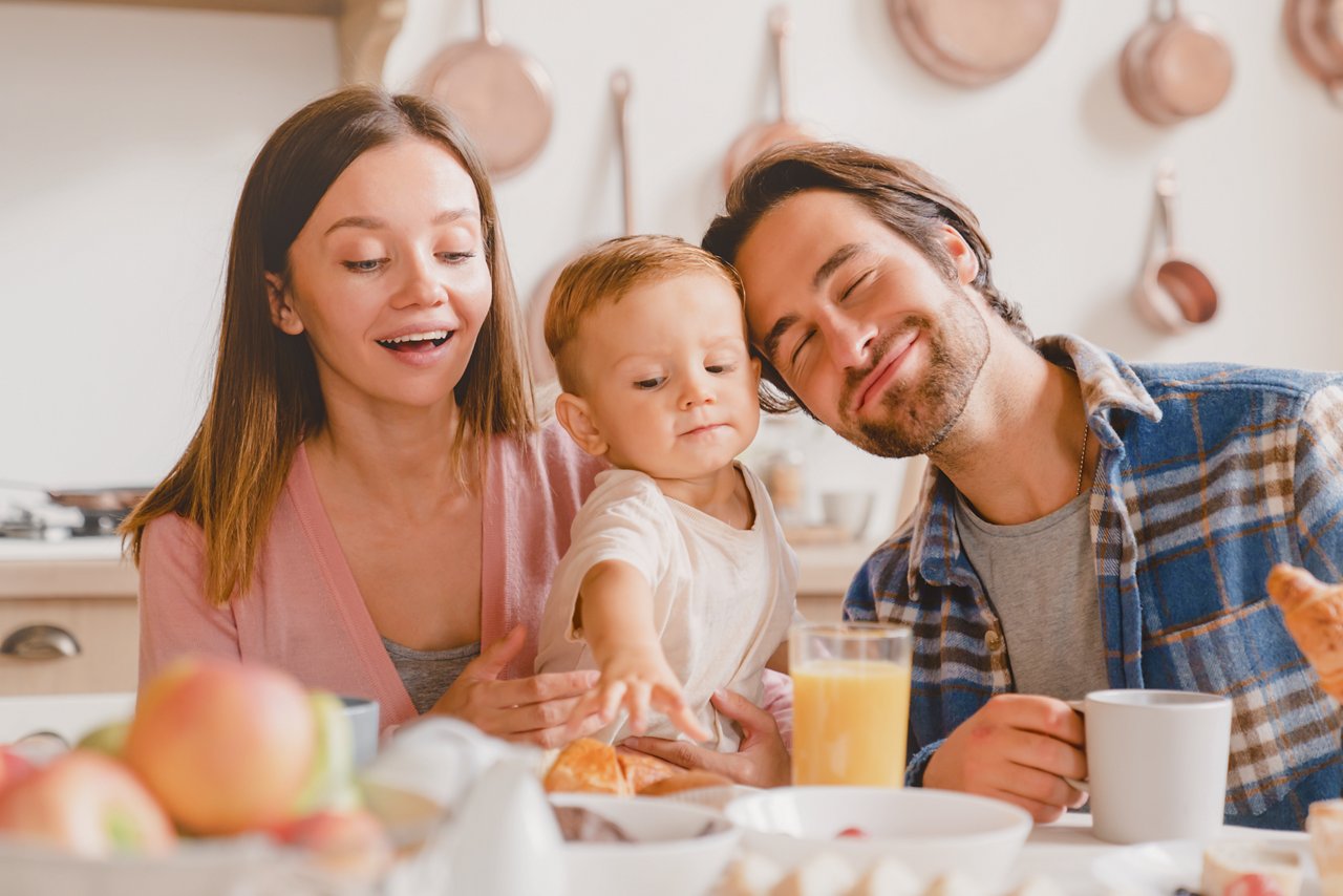 Family dinner lunch breakfast at home. Caucasian happy parents eating together, feeding small little kid child toddler new born baby infant at home kitchen. Adoption concept