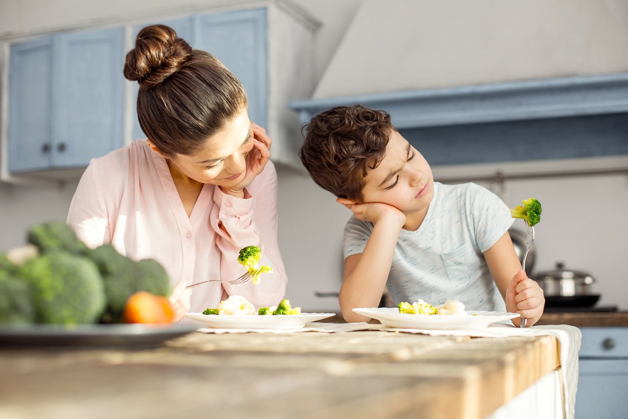Good food. Alert dark-haired young mom smiling and having healthy breakfast with her son and the boy looking at the vegetable on his fork
