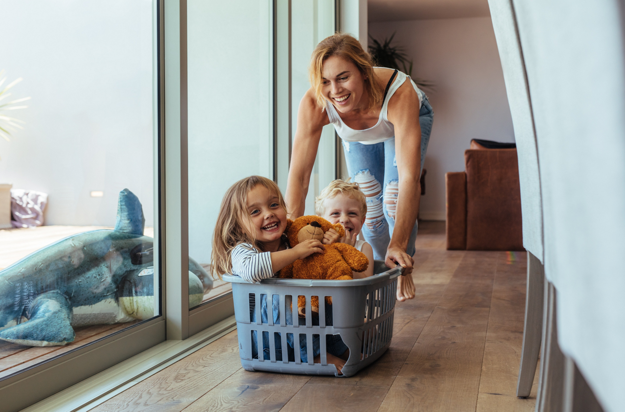 Eine Frau schiebt zwei lachende Kinder mit einem Teddy in einem Wäschekorb durch ein Wohnzimmer mit Holzboden.
