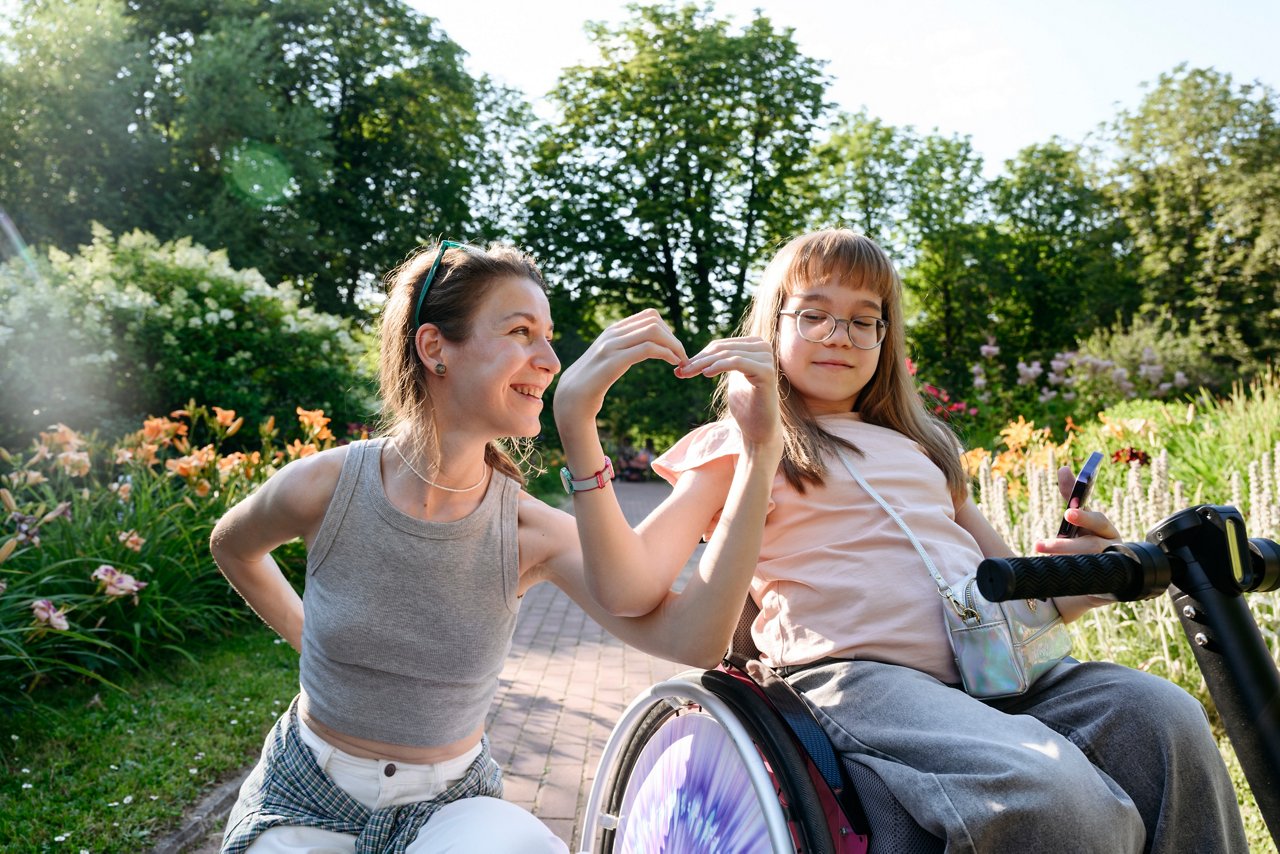 Smiling mother making heart shape with daughter sitting on wheelchair in park