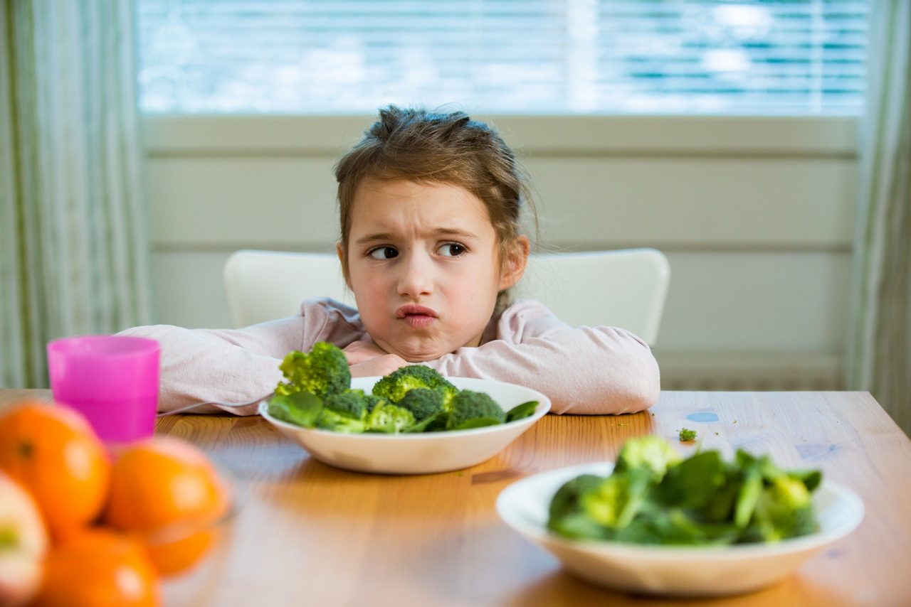 Cute girl eating spinach and broccoli at the table. Child doesn't want to eat, refuses eating, making faces. Healthy food concept.