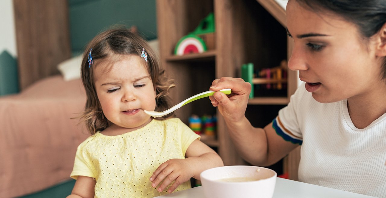 Eine Frau füttert ein kleines Mädchen mit Brei, beide sitzen an einem Tisch in einem Kinderzimmer mit Spielzeugregal.