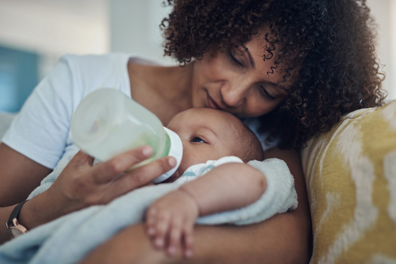 Eine Frau füttert ein Baby auf dem Sofa mit einem Fläschchen, das Baby ist in eine Decke gewickelt.