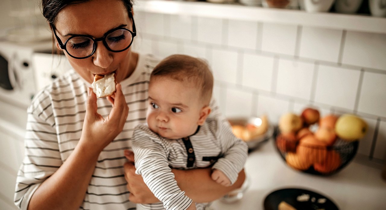 Mother eating in the kitchen while holding her baby