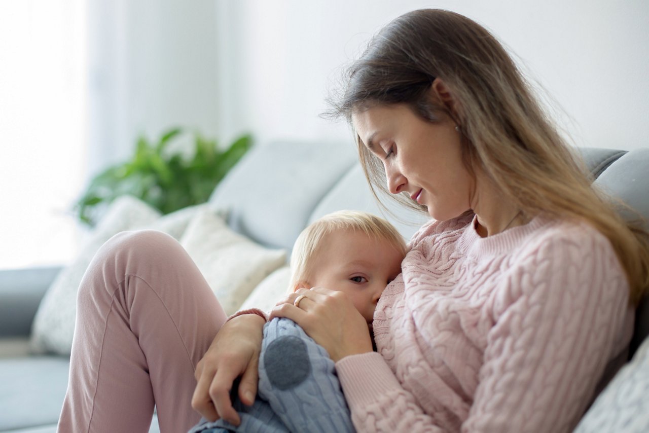 Young mother, holding her sick toddler boy, breastfeeding him at home, sunny living room