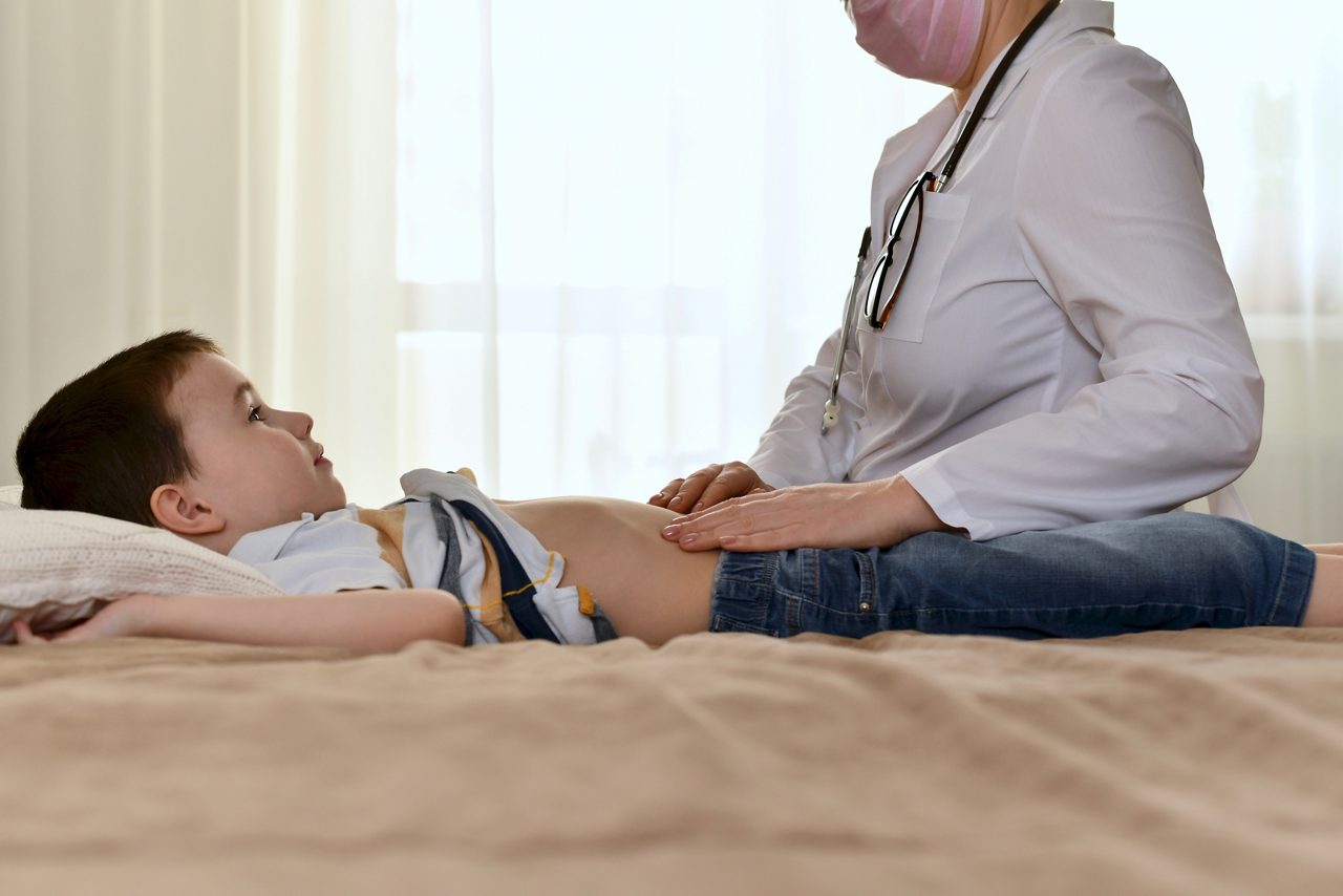 A doctor in a white coat checks the belly of a child. Sitting on the bed in the patient house. The boy looks at her with surprise.