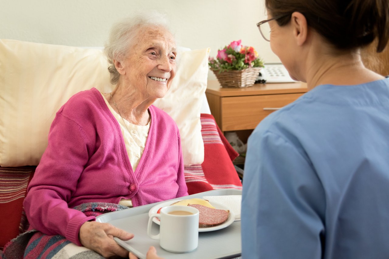 Ältere Schlaganfall-Patientin beim Essen lächelt Pflegerin an
